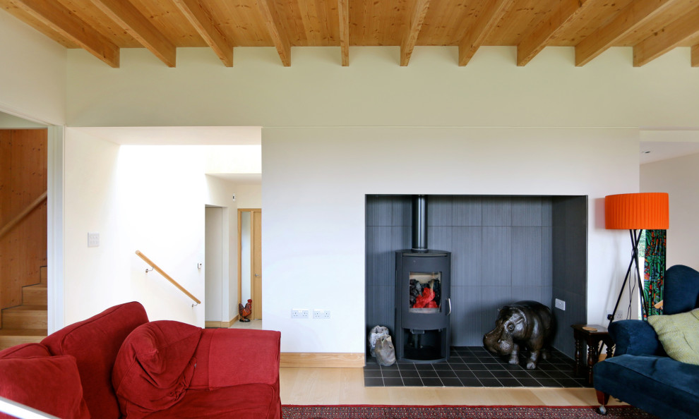 This is an example of a large contemporary enclosed living room in Devon with white walls, light hardwood floors, a wood stove, a tile fireplace surround, a freestanding tv, beige floor, exposed beam and panelled walls.