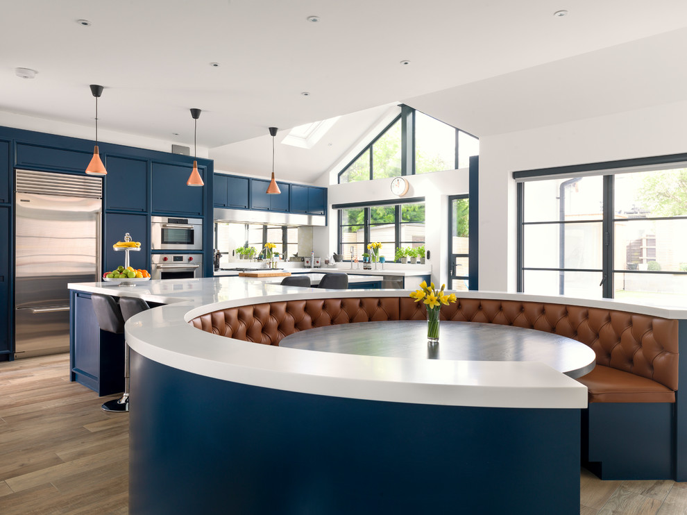 Transitional l-shaped eat-in kitchen in Other with shaker cabinets, blue cabinets, mirror splashback, stainless steel appliances, light hardwood floors, with island and white benchtop.