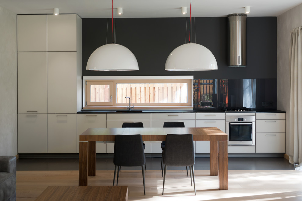Mid-sized contemporary single-wall eat-in kitchen in Moscow with an undermount sink, flat-panel cabinets, white cabinets, granite benchtops, black splashback, glass sheet splashback, stainless steel appliances, light hardwood floors, no island, beige floor and black benchtop.