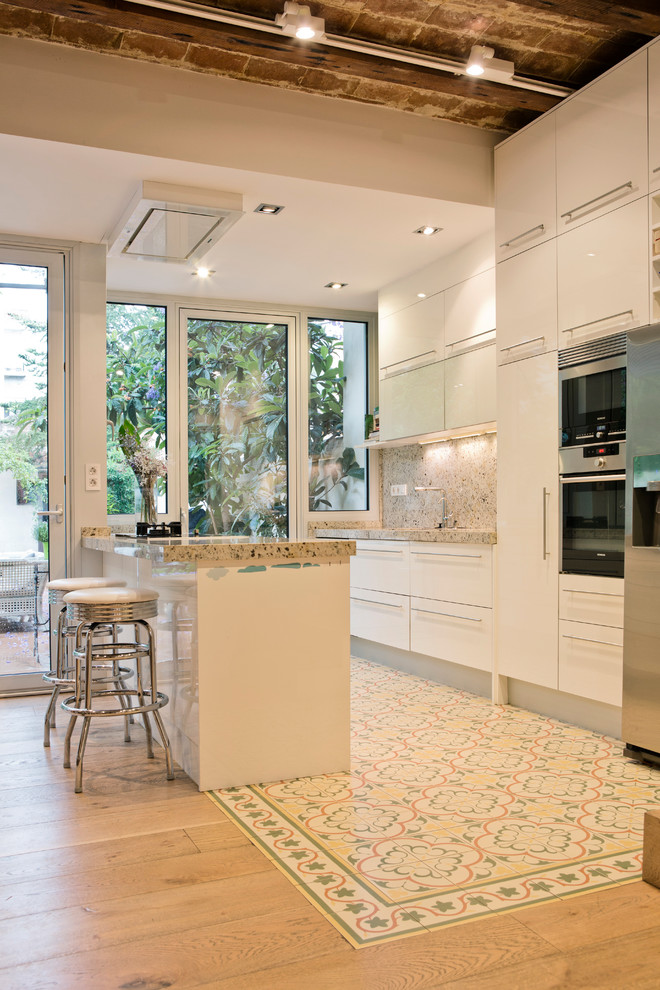 Inspiration for a contemporary single-wall open plan kitchen in Barcelona with flat-panel cabinets, white cabinets, stainless steel appliances, a peninsula, ceramic floors, beige splashback, stone slab splashback, multi-coloured floor and beige benchtop.