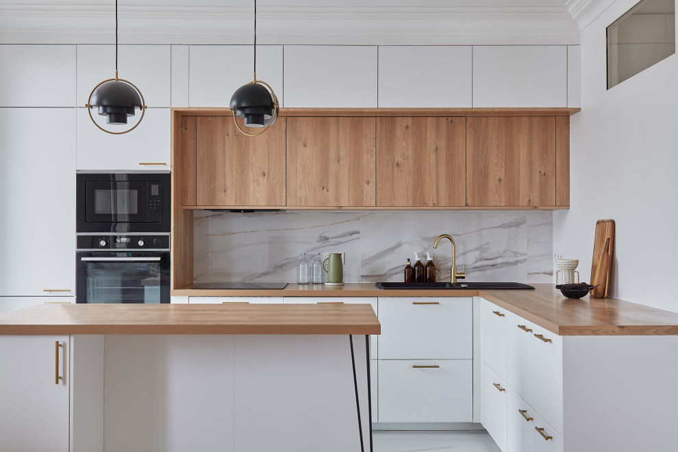 Inredning av ett minimalistiskt l-kök, med en nedsänkt diskho, släta luckor, träbänkskiva, grått stänkskydd, svarta vitvaror, en köksö, stänkskydd i marmor och marmorgolv