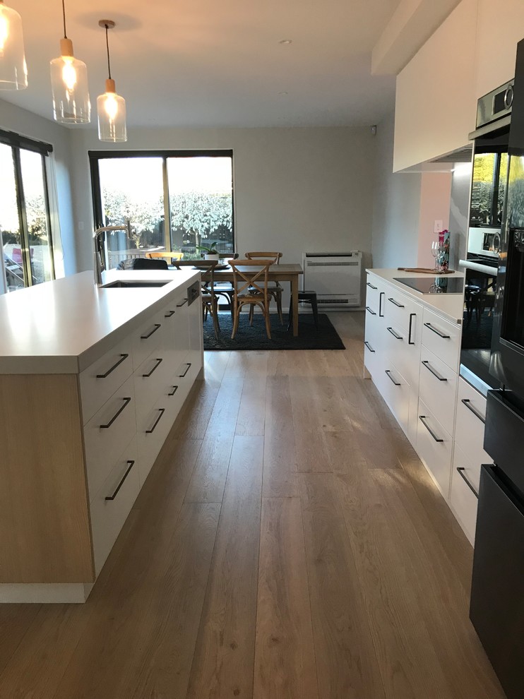 This is an example of a large contemporary galley open plan kitchen in Christchurch with a single-bowl sink, flat-panel cabinets, white cabinets, quartz benchtops, grey splashback, stone slab splashback, stainless steel appliances, medium hardwood floors, with island, brown floor and white benchtop.