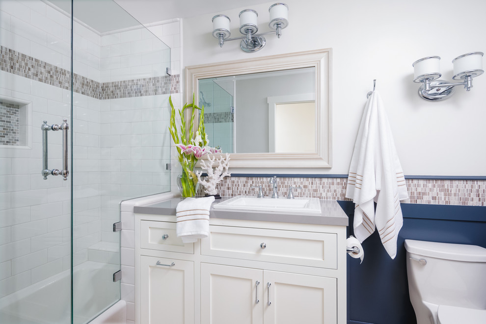 Mid-sized beach style 3/4 bathroom in San Diego with a drop-in sink, recessed-panel cabinets, white cabinets, solid surface benchtops, a drop-in tub, a shower/bathtub combo, a one-piece toilet, multi-coloured tile, mosaic tile, white walls and porcelain floors.