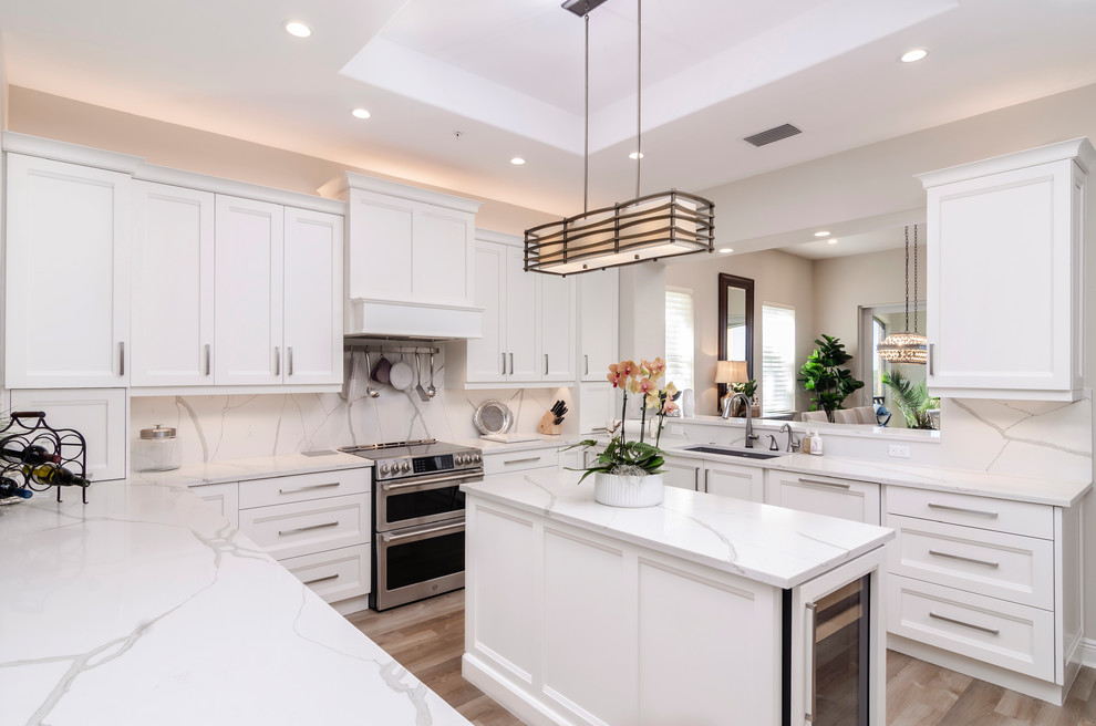 This is an example of a mid-sized transitional u-shaped open plan kitchen in Other with an undermount sink, white cabinets, stainless steel appliances, light hardwood floors, with island, white benchtop, shaker cabinets, quartz benchtops, white splashback, stone slab splashback and brown floor.