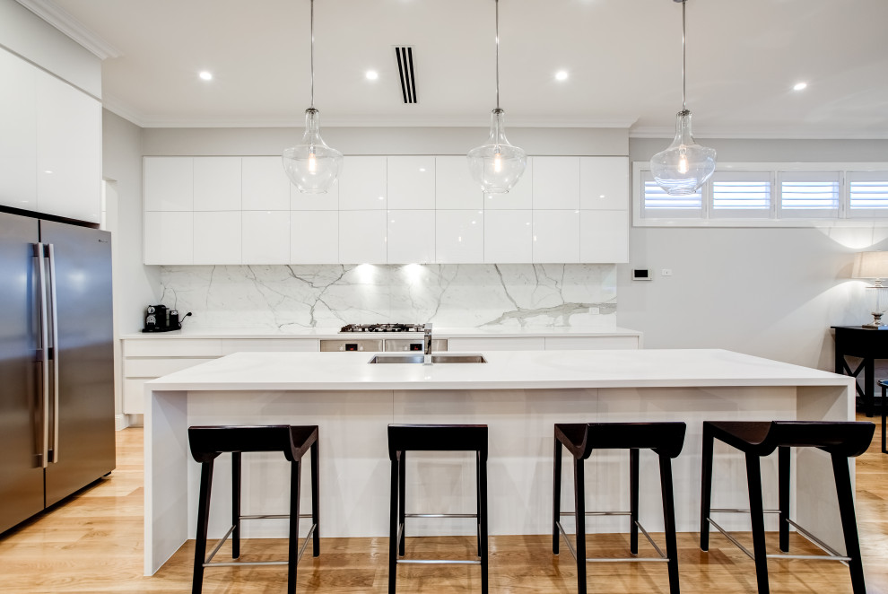 Photo of a large modern single-wall open plan kitchen in Brisbane with an undermount sink, flat-panel cabinets, white cabinets, white splashback, stainless steel appliances, with island, brown floor, white benchtop, laminate benchtops, marble splashback and light hardwood floors.