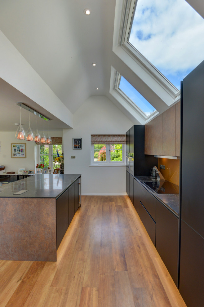 This is an example of a large modern l-shaped open plan kitchen in Sussex with an integrated sink, flat-panel cabinets, black cabinets, quartzite benchtops, brown splashback, glass sheet splashback, black appliances, laminate floors, with island, brown floor, black benchtop and vaulted.