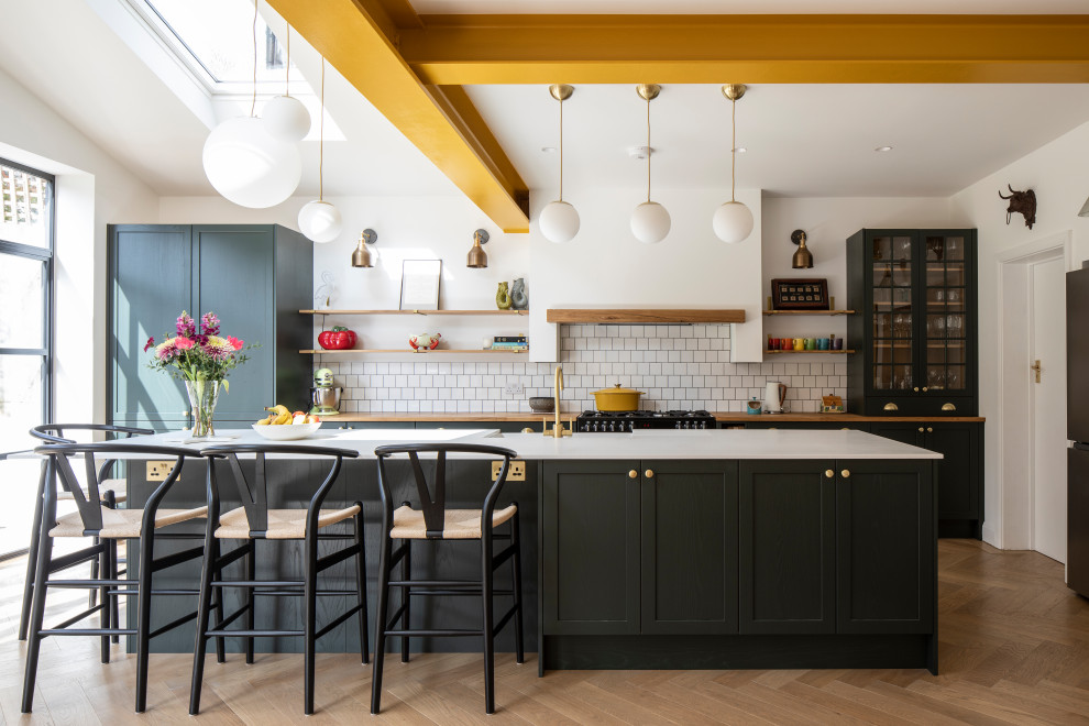 Photo of a transitional galley kitchen in London with a farmhouse sink, shaker cabinets, black cabinets, wood benchtops, white splashback, black appliances, medium hardwood floors, with island, brown floor, brown benchtop and exposed beam.