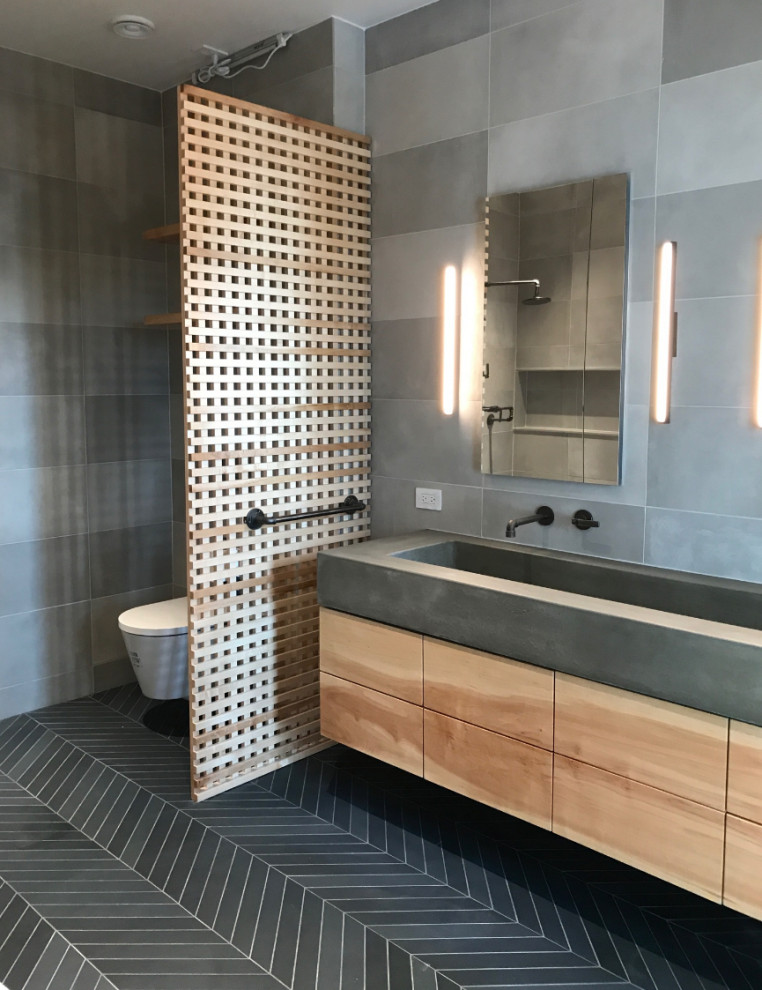 Industriell inredning av ett mellanstort grå grått en-suite badrum, med släta luckor, skåp i ljust trä, ett fristående badkar, en öppen dusch, en vägghängd toalettstol, grå kakel, porslinskakel, grå väggar, klinkergolv i porslin, ett integrerad handfat, bänkskiva i betong, svart golv och med dusch som är öppen