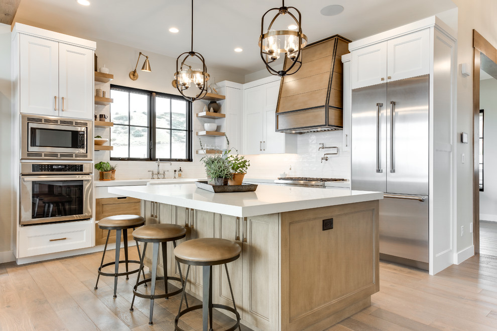 This is an example of a country l-shaped eat-in kitchen in Salt Lake City with white splashback, stainless steel appliances, with island, white benchtop, a farmhouse sink, recessed-panel cabinets, medium wood cabinets, medium hardwood floors and brown floor.