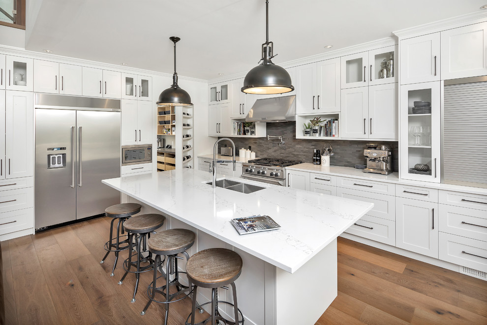 Mid-sized transitional l-shaped medium tone wood floor and brown floor kitchen photo with a double-bowl sink, shaker cabinets, white cabinets, gray backsplash, stainless steel appliances and an island