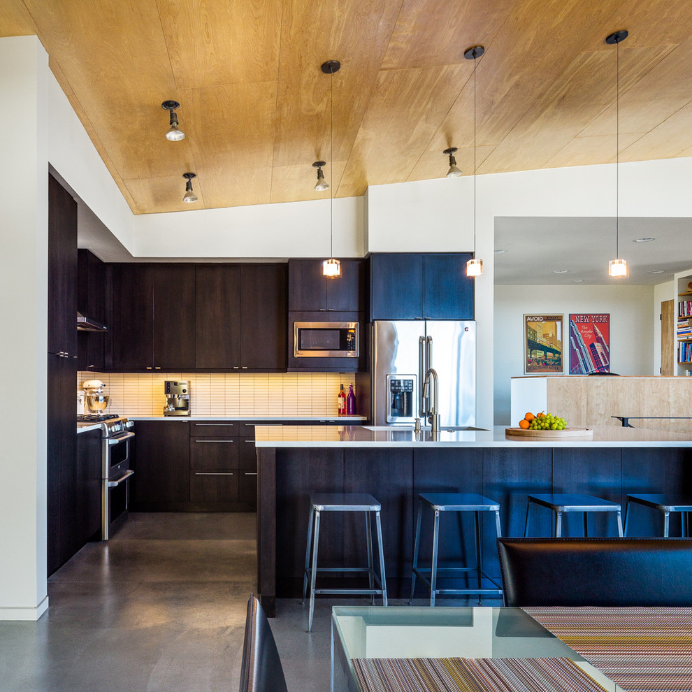 Mid-sized contemporary l-shaped open plan kitchen in Seattle with flat-panel cabinets, black cabinets, white splashback, glass tile splashback, stainless steel appliances, concrete floors, with island, grey floor and white benchtop.