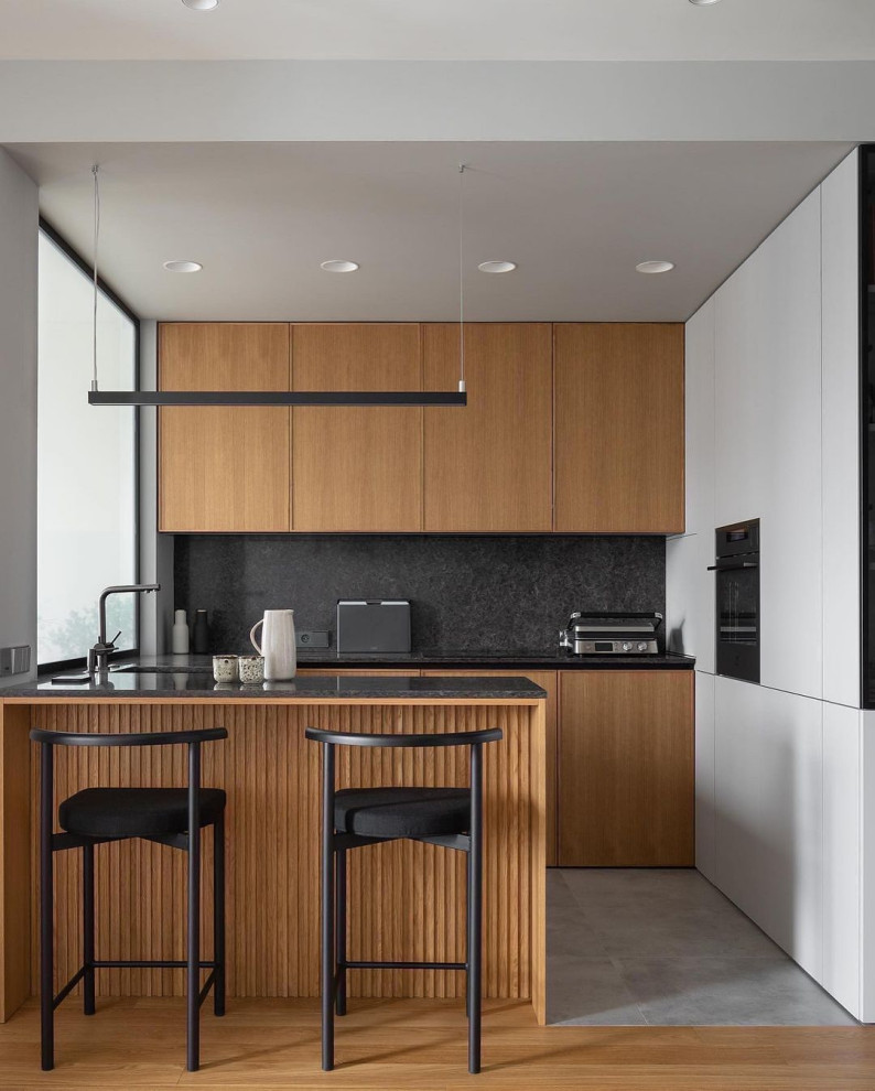 Foto di una cucina ad U design con ante lisce, ante in legno chiaro, paraspruzzi nero, elettrodomestici neri, penisola, pavimento grigio e top nero