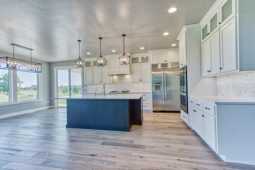 Example of a large transitional l-shaped light wood floor and brown floor eat-in kitchen design with an undermount sink, shaker cabinets, white cabinets, quartz countertops, white backsplash, ceramic backsplash, stainless steel appliances, an island and white countertops