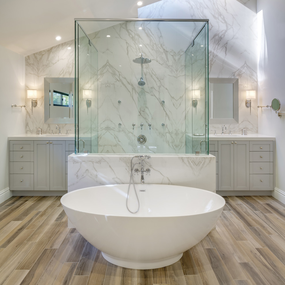 This is an example of a contemporary master wet room bathroom in Essex with recessed-panel cabinets, grey cabinets, a freestanding tub, white tile, marble, white walls, light hardwood floors, an integrated sink, marble benchtops, brown floor and a hinged shower door.