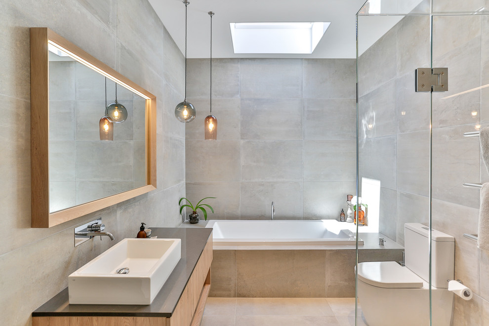Inspiration for a mid-sized contemporary 3/4 bathroom in Auckland with light wood cabinets, a one-piece toilet, gray tile, ceramic tile, grey walls, engineered quartz benchtops, black benchtops, flat-panel cabinets, a drop-in tub, a vessel sink and beige floor.