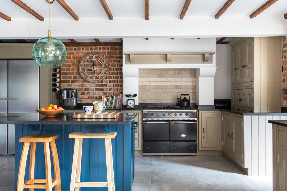 Mid-sized country l-shaped kitchen with blue cabinets, with island, grey floor, black benchtop, recessed-panel cabinets, beige splashback, brick splashback and black appliances.