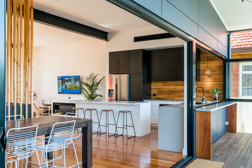 Photo of a mid-sized contemporary l-shaped open plan kitchen in Sydney with quartz benchtops, black appliances, light hardwood floors, with island, white benchtop, flat-panel cabinets, black cabinets, brown splashback, timber splashback and brown floor.