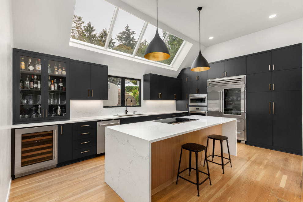 Contemporary l-shaped kitchen in Portland with a submerged sink, flat-panel cabinets, black cabinets, engineered stone countertops, white splashback, engineered quartz splashback, stainless steel appliances, light hardwood flooring, an island, white worktops and a vaulted ceiling.