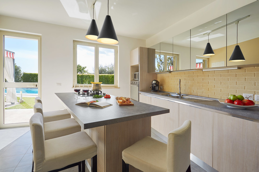 Design ideas for a contemporary galley open plan kitchen in Other with with island, light wood cabinets, brick splashback, porcelain floors, grey floor, a drop-in sink, flat-panel cabinets, yellow splashback, stainless steel appliances and grey benchtop.