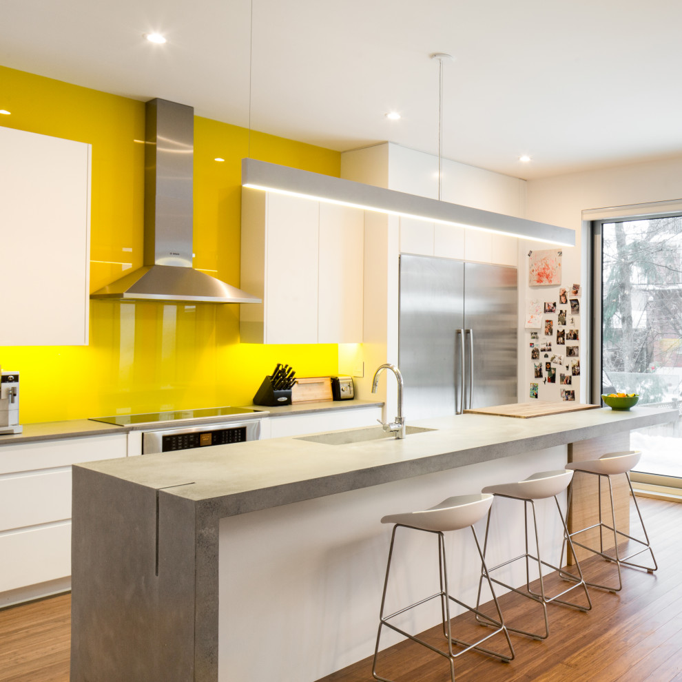 Photo of a large contemporary galley kitchen in Ottawa with an undermount sink, flat-panel cabinets, white cabinets, concrete benchtops, yellow splashback, glass sheet splashback, stainless steel appliances, medium hardwood floors, with island, brown floor and grey benchtop.