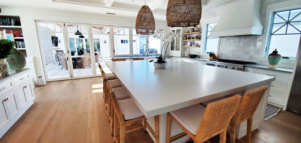 Photo of an expansive beach style u-shaped kitchen in San Diego with a farmhouse sink, recessed-panel cabinets, white cabinets, quartz benchtops, white splashback, ceramic splashback, stainless steel appliances, medium hardwood floors, with island, grey benchtop and coffered.