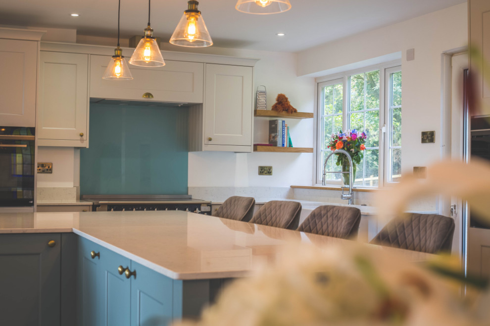 Design ideas for a large contemporary l-shaped open plan kitchen in Devon with a farmhouse sink, shaker cabinets, turquoise cabinets, quartzite benchtops, blue splashback, glass sheet splashback, black appliances, light hardwood floors, with island, brown floor and white benchtop.