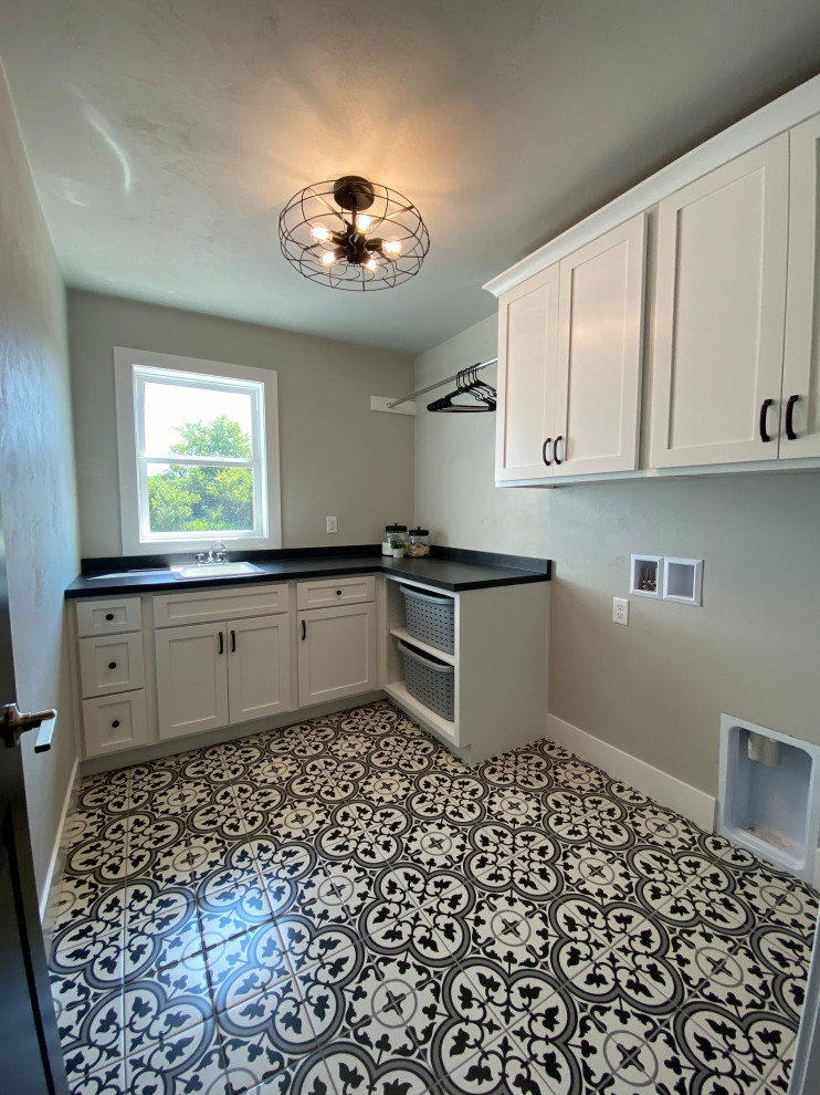 Large l-shaped utility room in Other with an utility sink, shaker cabinets, white cabinets, beige walls, terra-cotta floors, a side-by-side washer and dryer, multi-coloured floor and black benchtop.