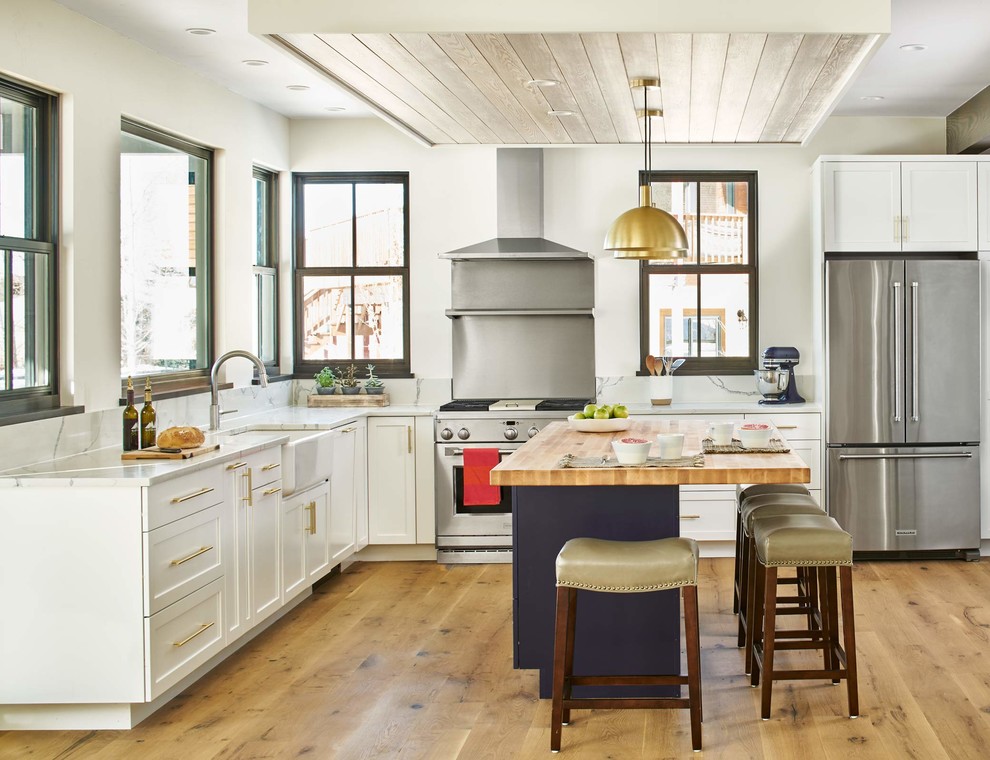 Design ideas for a country l-shaped kitchen in Denver with a farmhouse sink, shaker cabinets, white cabinets, wood benchtops, metallic splashback, stainless steel appliances, light hardwood floors and with island.
