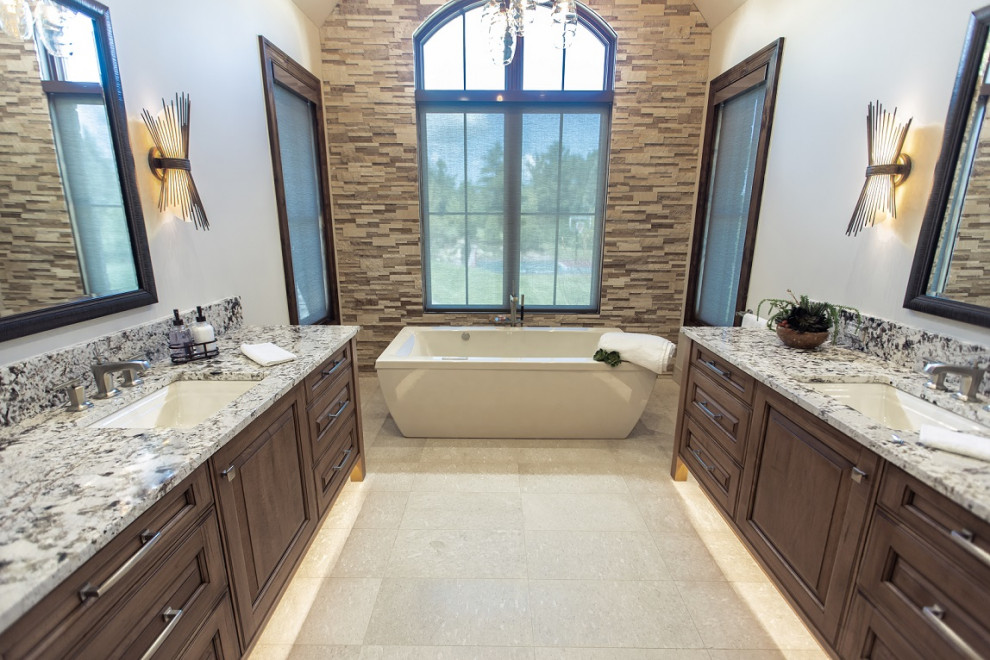 This is an example of a large transitional master bathroom in Chicago with raised-panel cabinets, medium wood cabinets, a freestanding tub, gray tile, travertine, limestone floors, an undermount sink, granite benchtops, beige floor and multi-coloured benchtops.
