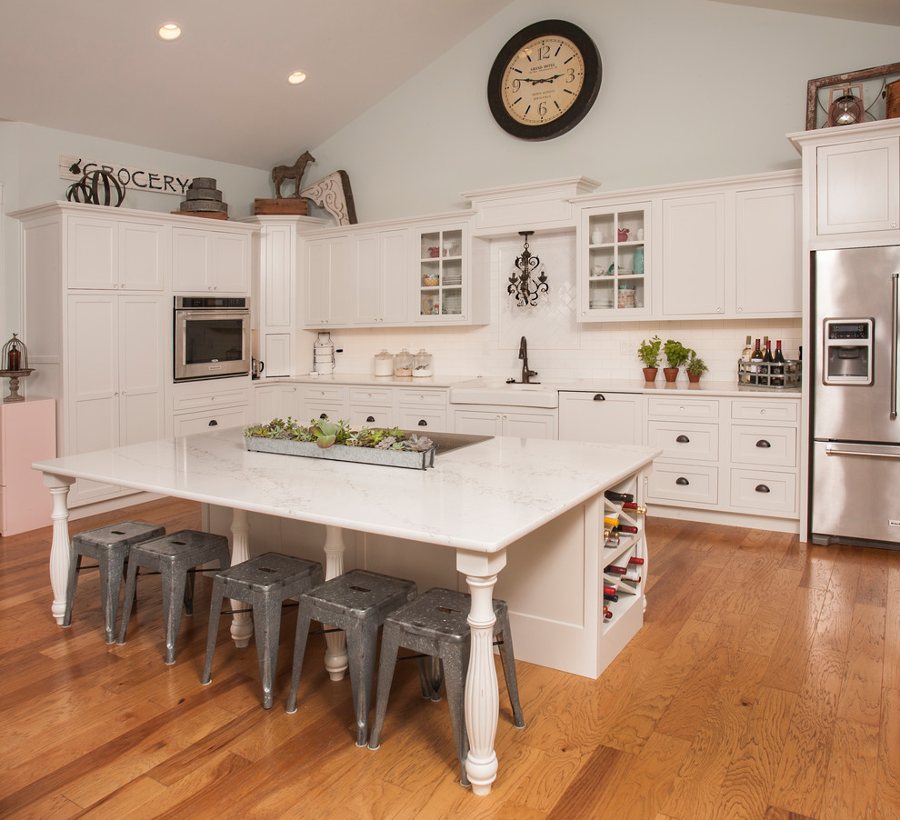 Photo of a country l-shaped kitchen in Seattle with a farmhouse sink, shaker cabinets, white cabinets, white splashback, subway tile splashback, stainless steel appliances, medium hardwood floors, with island and brown floor.