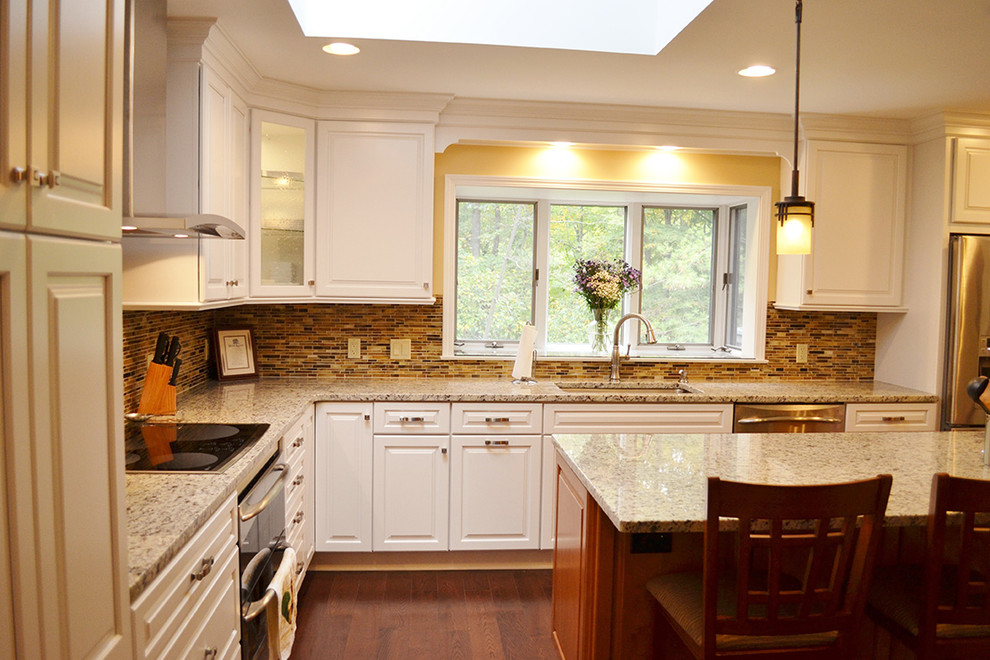 Amerikansk inredning av ett stort kök, med en undermonterad diskho, luckor med infälld panel, vita skåp, granitbänkskiva, flerfärgad stänkskydd, stänkskydd i glaskakel, rostfria vitvaror, mörkt trägolv och en köksö
