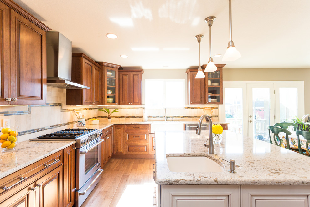 Example of a large transitional l-shaped medium tone wood floor and brown floor eat-in kitchen design in Denver with a farmhouse sink, raised-panel cabinets, medium tone wood cabinets, granite countertops, beige backsplash, stone tile backsplash, stainless steel appliances and an island