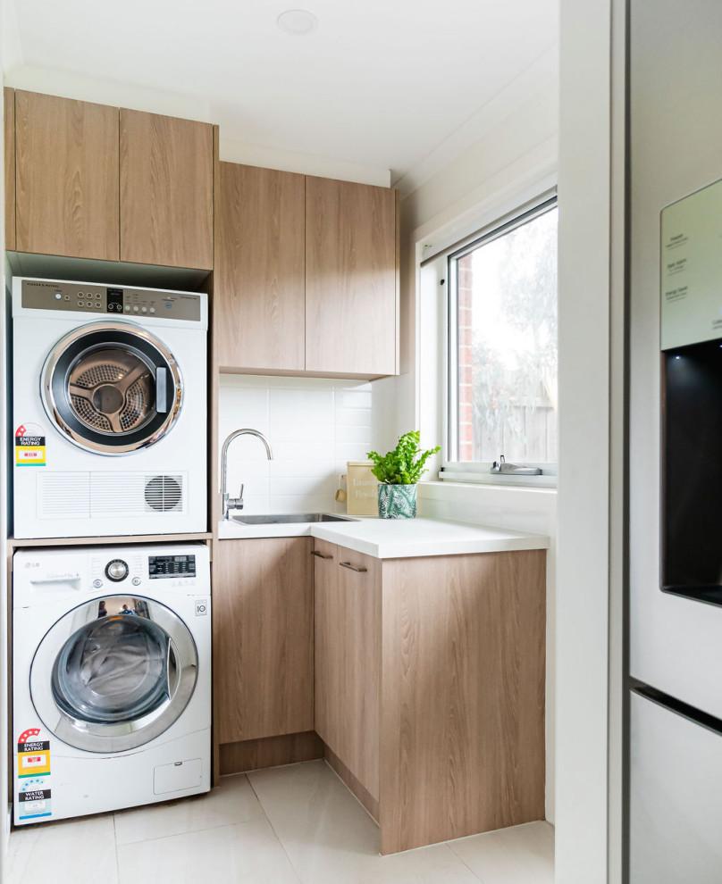 Inspiration for a small modern l-shaped dedicated laundry room in Melbourne with flat-panel cabinets, quartz benchtops, blue splashback, ceramic splashback, porcelain floors, beige floor, white benchtop, a drop-in sink, light wood cabinets, white walls and a stacked washer and dryer.