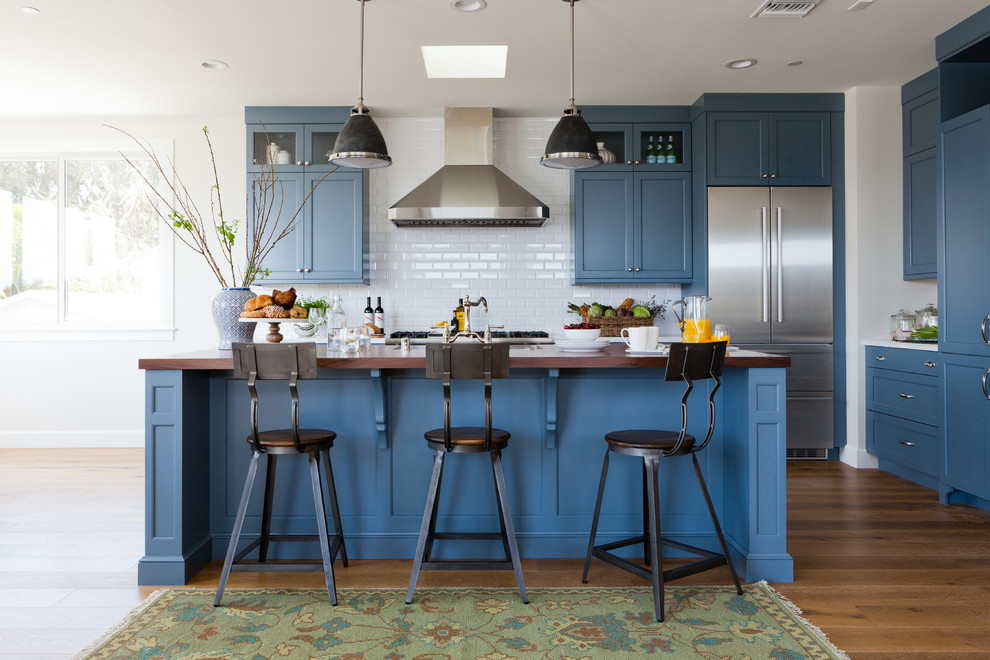 Mid-sized beach style l-shaped open plan kitchen in Los Angeles with shaker cabinets, blue cabinets, wood benchtops, white splashback, ceramic splashback, stainless steel appliances, medium hardwood floors and with island.