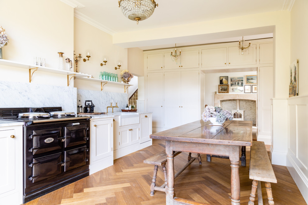 Inspiration for a large traditional eat-in kitchen in Other with a farmhouse sink, shaker cabinets, beige cabinets, marble benchtops, white splashback, marble splashback, black appliances, medium hardwood floors, no island, brown floor and white benchtop.
