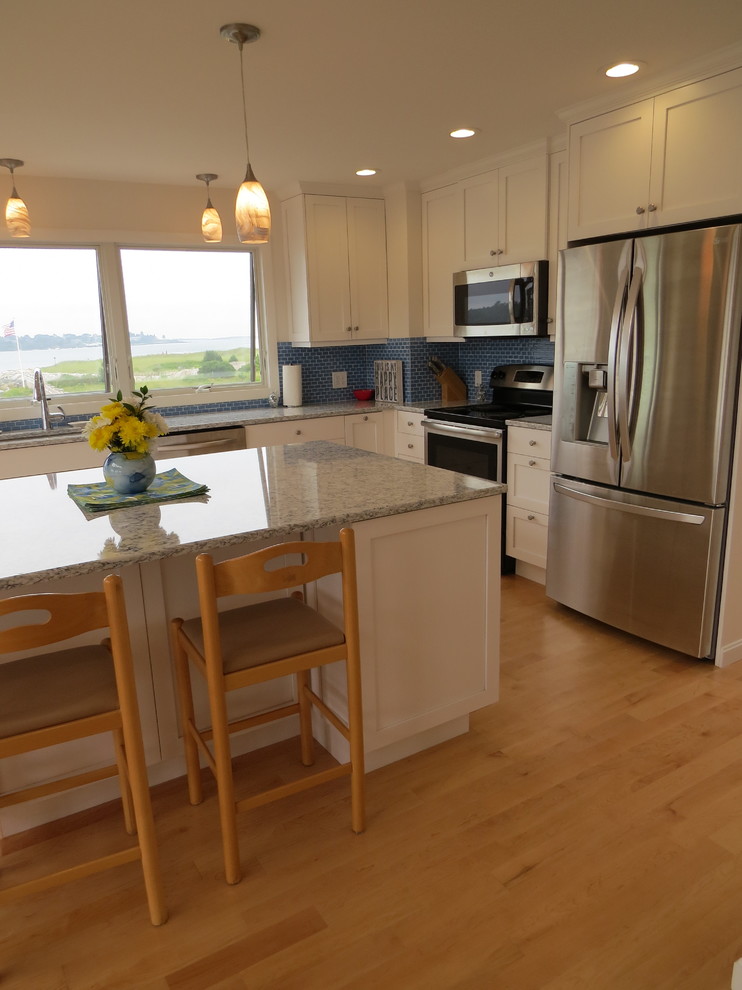 This is an example of a mid-sized beach style l-shaped open plan kitchen in Portland Maine with an undermount sink, shaker cabinets, white cabinets, quartz benchtops, blue splashback, glass tile splashback, stainless steel appliances, light hardwood floors and with island.