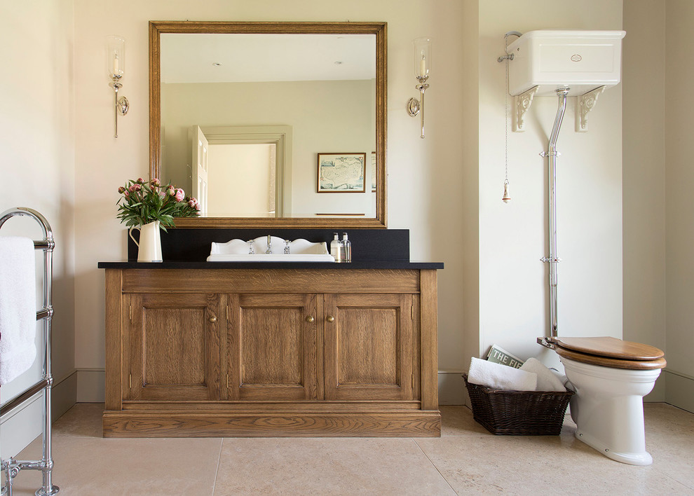 Design ideas for a mid-sized traditional bathroom in Gloucestershire with recessed-panel cabinets, medium wood cabinets, a one-piece toilet, beige walls, a drop-in sink, beige floor and black benchtops.