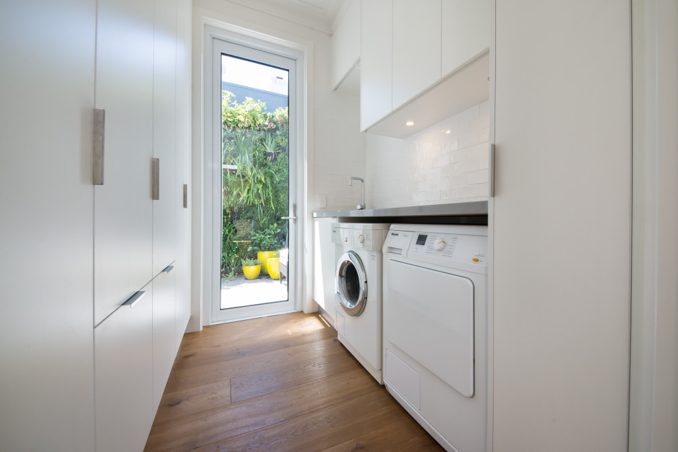 Expansive beach style single-wall dedicated laundry room in Melbourne with an undermount sink, flat-panel cabinets, white cabinets, quartz benchtops, white walls, medium hardwood floors, a side-by-side washer and dryer and brown floor.