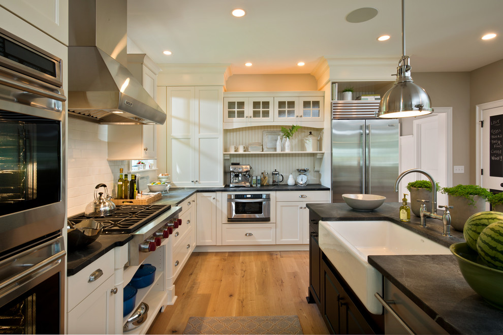 This is an example of a mid-sized traditional l-shaped separate kitchen in Boston with a farmhouse sink, white splashback, subway tile splashback, stainless steel appliances, shaker cabinets, white cabinets, solid surface benchtops, light hardwood floors and with island.