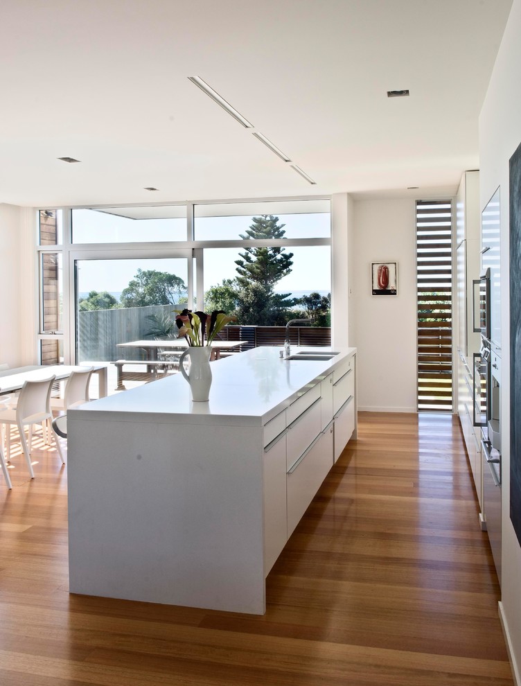 Inspiration for a contemporary eat-in kitchen in Wellington with a double-bowl sink, white cabinets, quartz benchtops, grey splashback, glass sheet splashback, stainless steel appliances, light hardwood floors and with island.