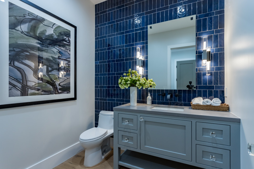 Design ideas for a mid-sized transitional powder room in Los Angeles with shaker cabinets, blue cabinets, a one-piece toilet, blue tile, ceramic tile, grey walls, light hardwood floors, an undermount sink, beige floor, grey benchtops, a built-in vanity and recessed.