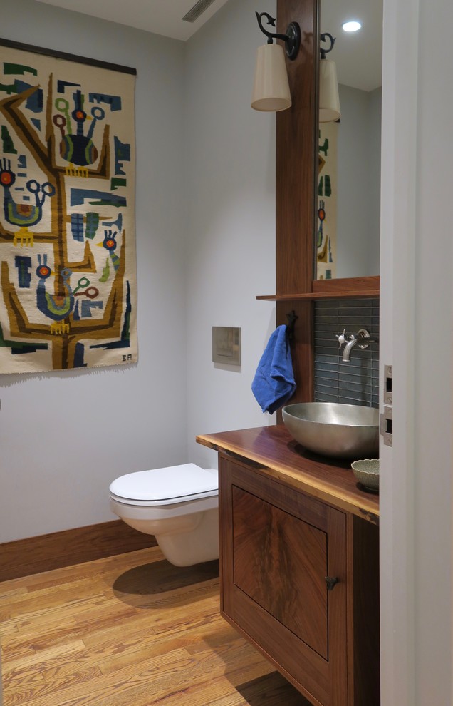 This is an example of a contemporary powder room in New York with furniture-like cabinets, dark wood cabinets, a wall-mount toilet, blue tile, glass tile, grey walls, medium hardwood floors and a vessel sink.