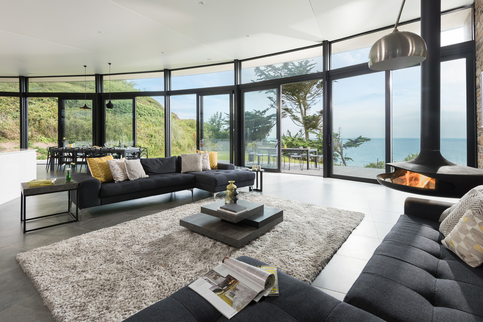 Inspiration for a large contemporary open concept living room in Cornwall with grey walls, porcelain floors, a hanging fireplace, a stone fireplace surround, a wall-mounted tv and grey floor.