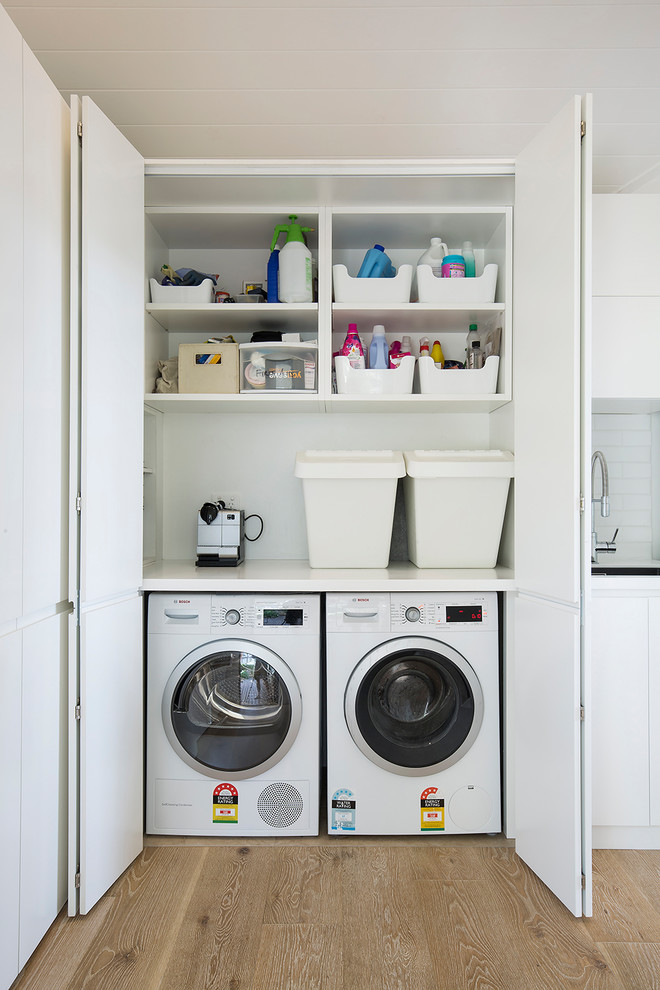 Photo of a contemporary single-wall laundry cupboard in Sydney with an undermount sink, flat-panel cabinets, white cabinets, white walls, medium hardwood floors, a side-by-side washer and dryer, brown floor and white benchtop.