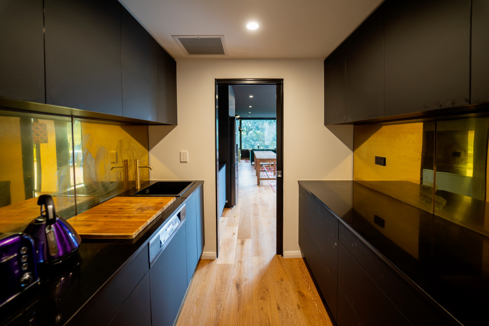 Photo of a mid-sized asian galley separate kitchen in Sydney with a drop-in sink, flat-panel cabinets, black cabinets, quartz benchtops, metallic splashback, metal splashback, black appliances, light hardwood floors, no island, brown floor and black benchtop.