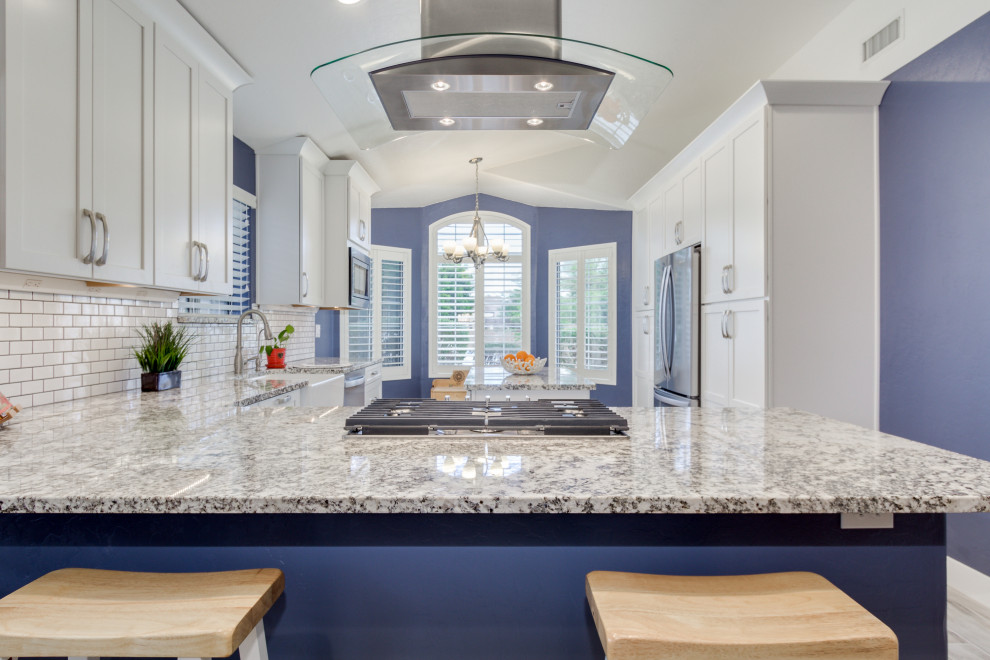 Beach style porcelain tile and gray floor kitchen photo with a farmhouse sink, shaker cabinets, white cabinets, granite countertops, white backsplash, subway tile backsplash, stainless steel appliances, an island and multicolored countertops