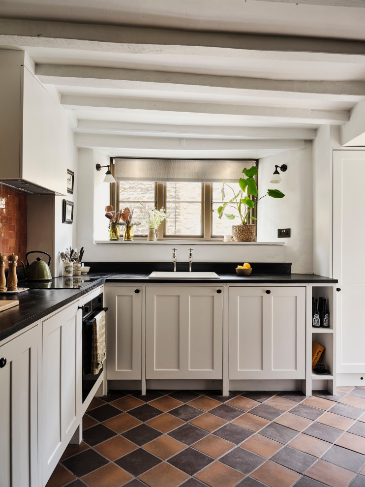 Small country l-shaped separate kitchen in Oxfordshire with shaker cabinets, white cabinets, wood benchtops, brown splashback, mosaic tile splashback, black appliances, ceramic floors, no island, multi-coloured floor, brown benchtop and exposed beam.