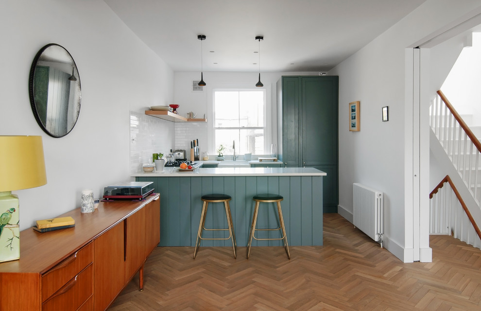 Inspiration for a transitional u-shaped eat-in kitchen in London with a farmhouse sink, shaker cabinets, blue cabinets, white splashback, subway tile splashback, medium hardwood floors, a peninsula, brown floor and white benchtop.