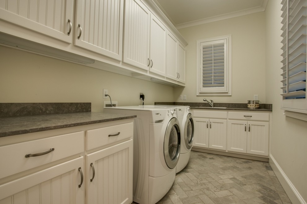 This is an example of a mid-sized traditional l-shaped dedicated laundry room in Dallas with a drop-in sink, recessed-panel cabinets, white cabinets, quartz benchtops, beige walls, brick floors, a side-by-side washer and dryer and beige floor.