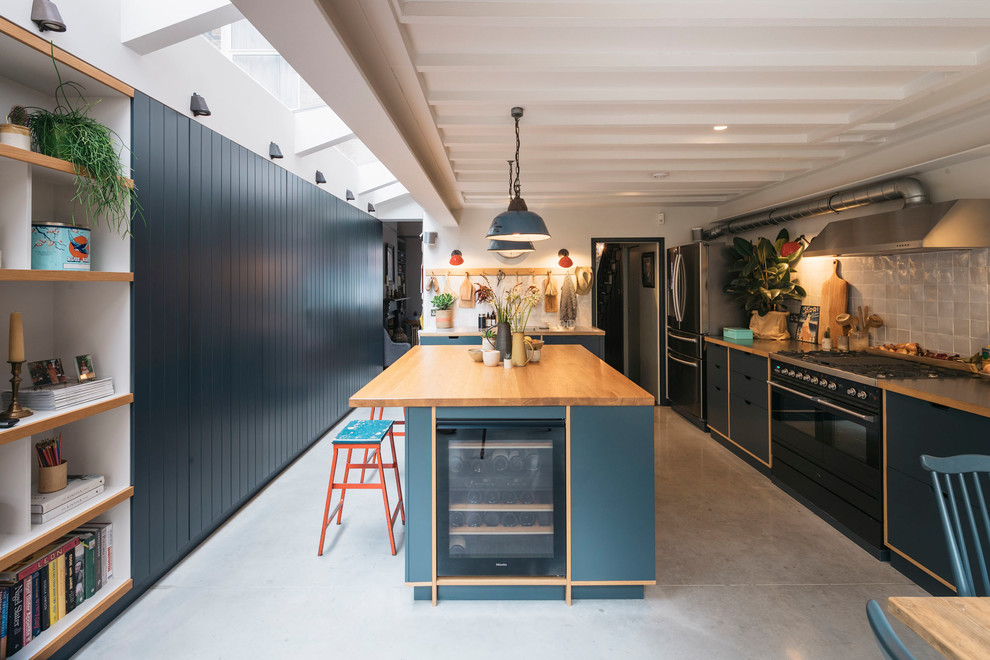 This is an example of a contemporary galley eat-in kitchen in London with flat-panel cabinets, blue cabinets, wood benchtops, beige splashback, porcelain splashback, concrete floors, with island, grey floor and black appliances.